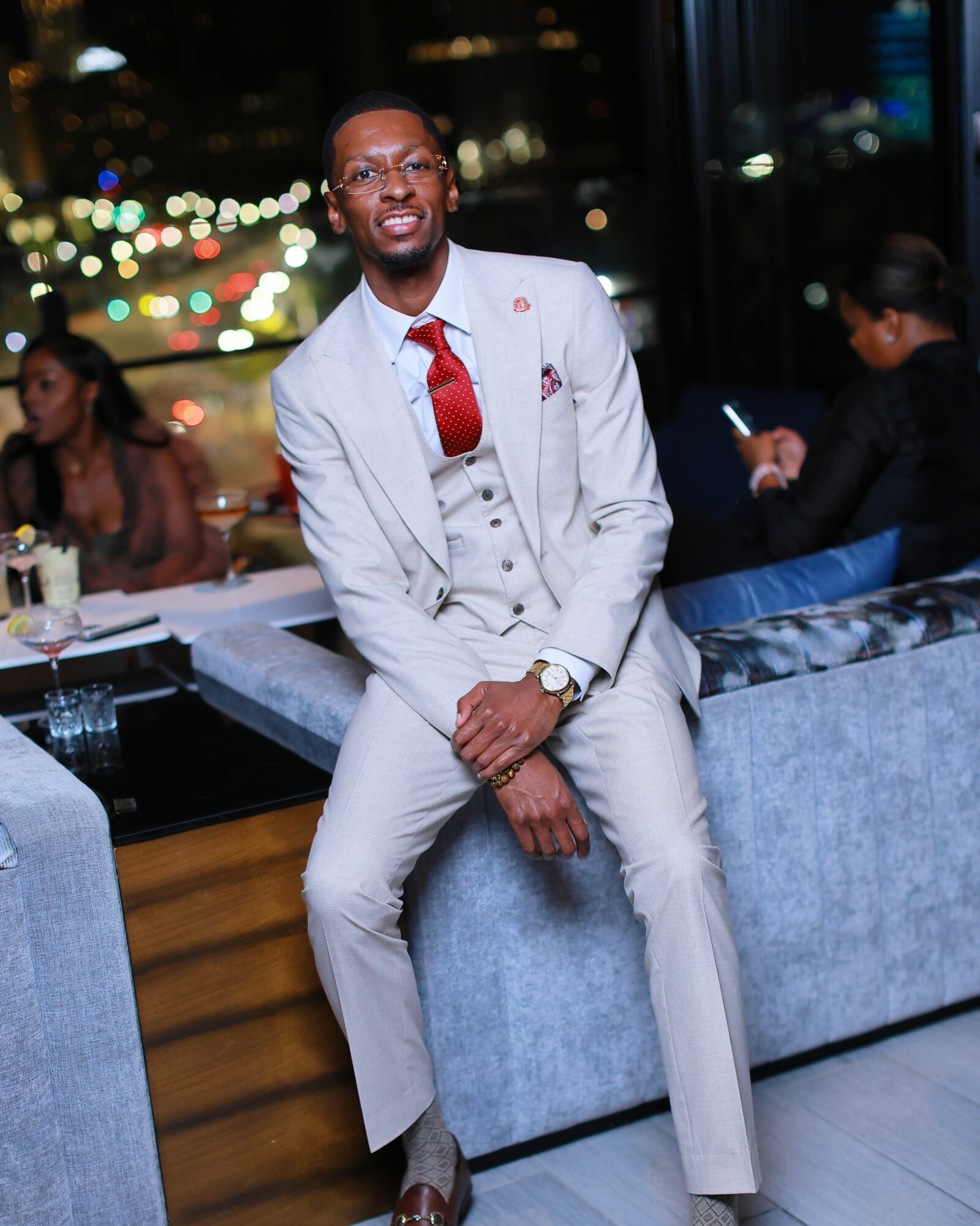 Man in light gray suit and red tie sitting on a blue sofa in a restaurant or lounge with city lights outside.