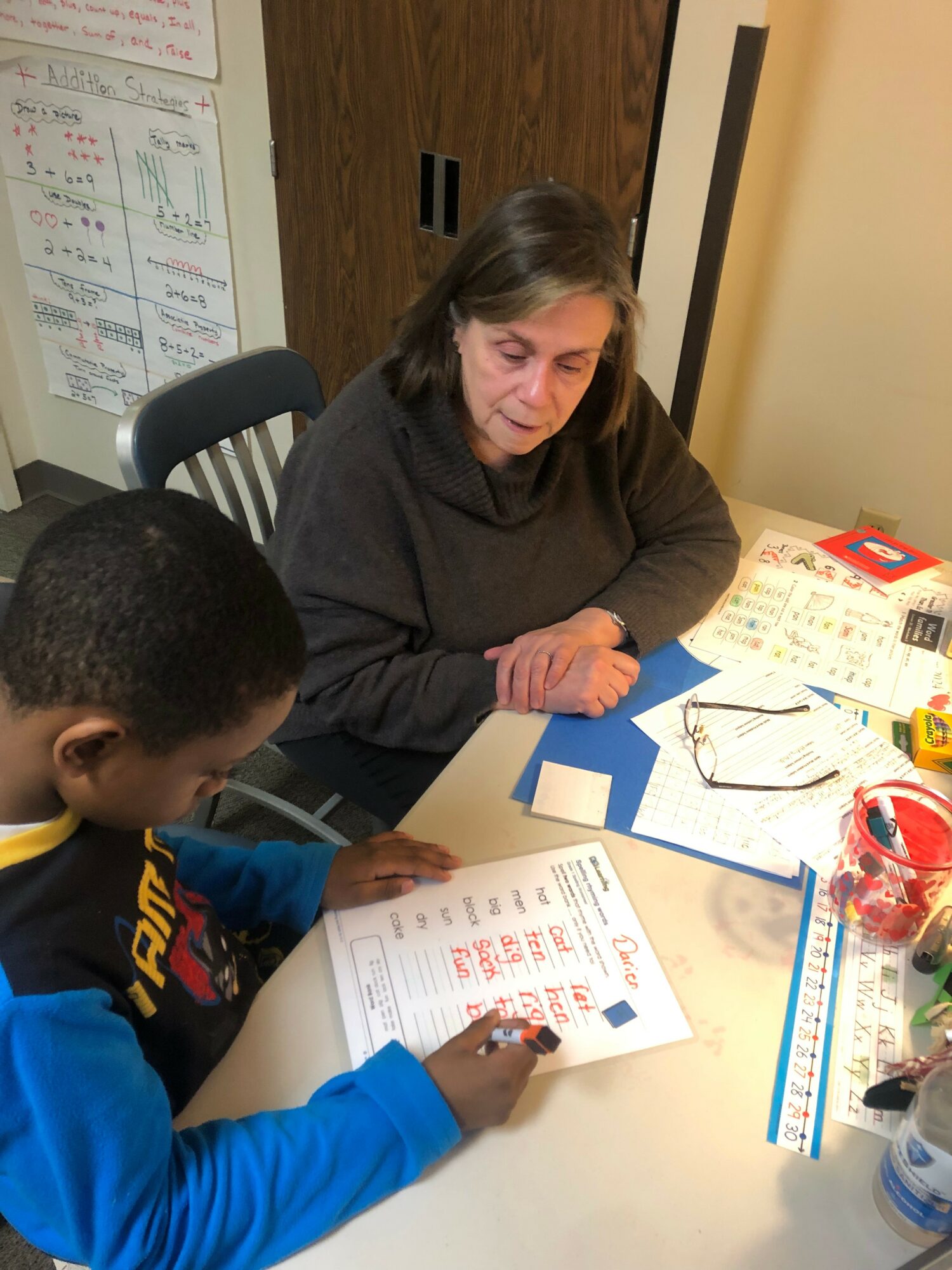 Woman and child sitting at a table with papers, books, and writing tools, engaging in an activity.