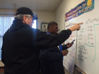 Two men standing in front of a whiteboard, one pointing at writing on the board, the other holding a marker.