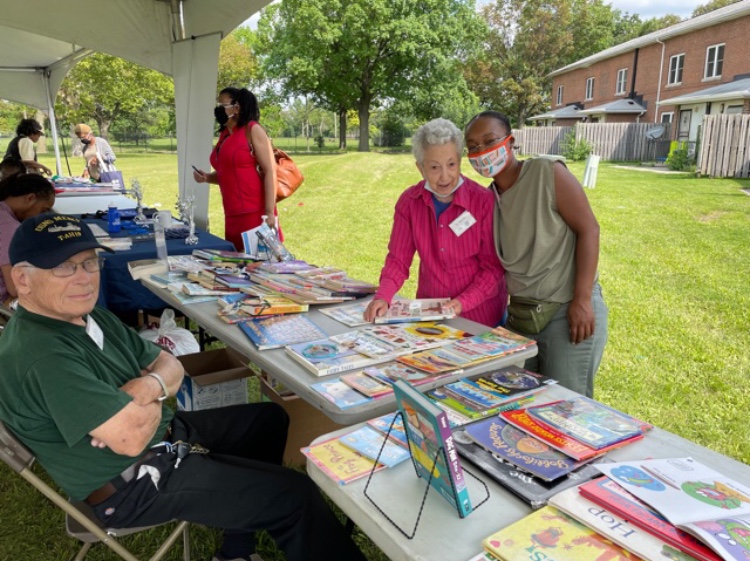 Two women stand behind a table filled with books outdoors, with a man sitting nearby and other people in the background.
