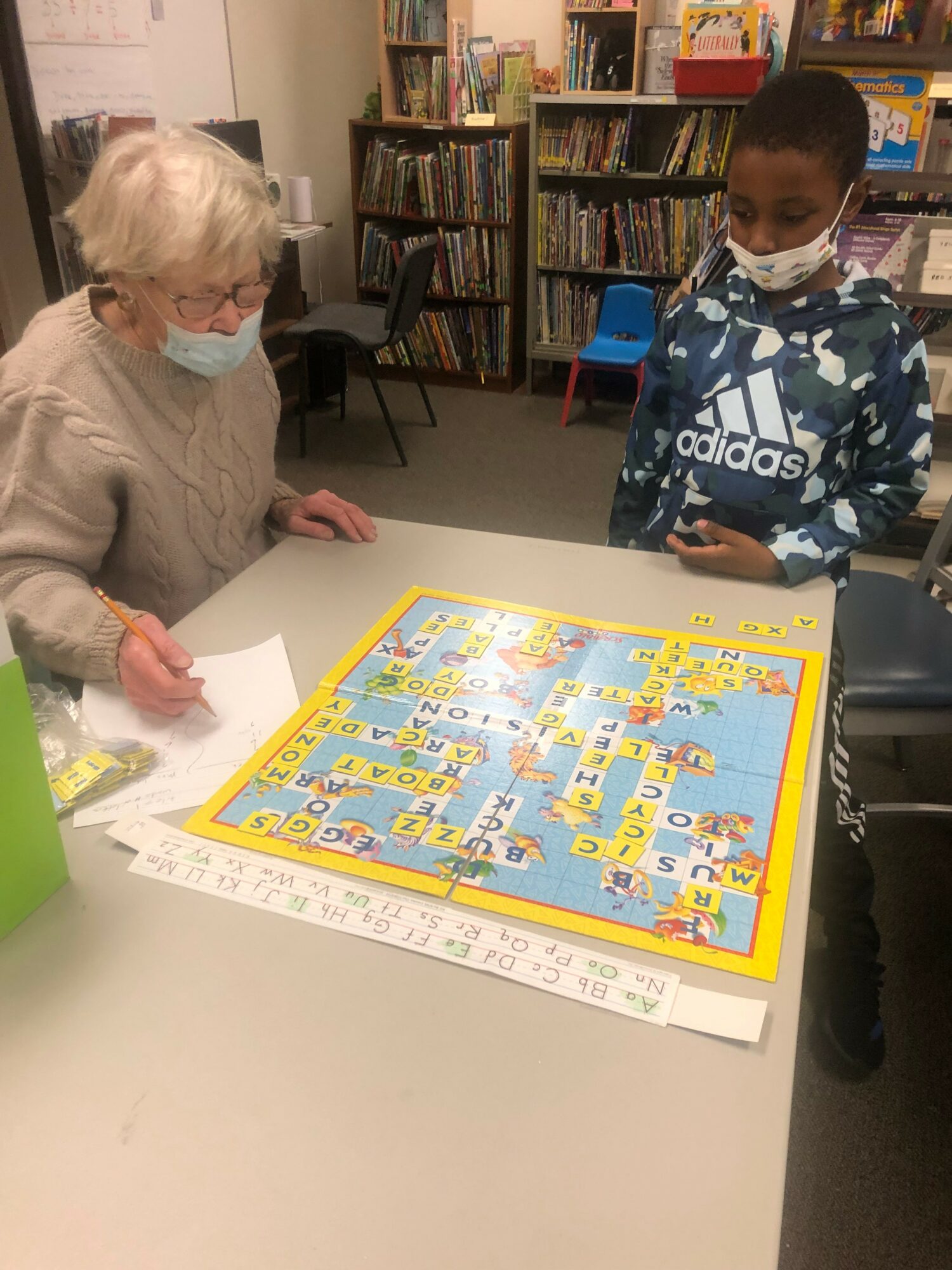 An elderly woman and a young boy wearing a face mask play a board game on a table in a library.