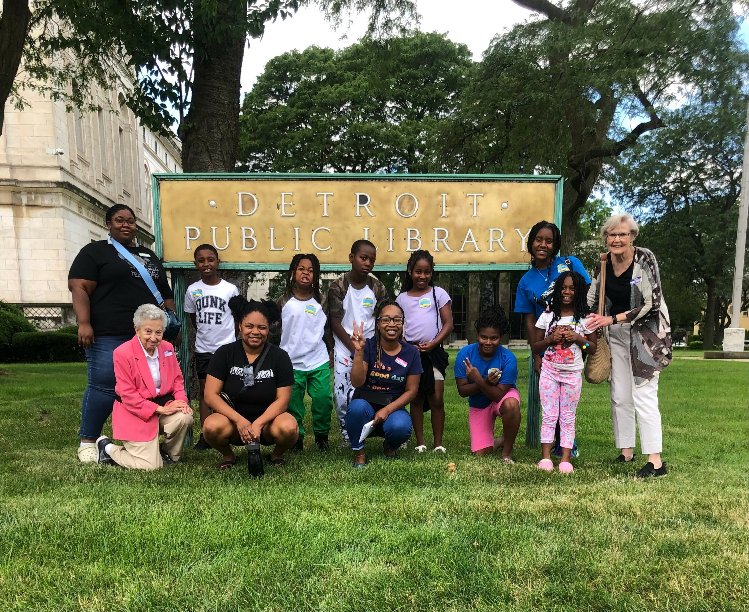 Group of children and adults posing in front of Detroit Public Library sign outdoors.