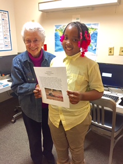 An elderly woman and a young girl stand together indoors, holding a paper and smiling at the camera.