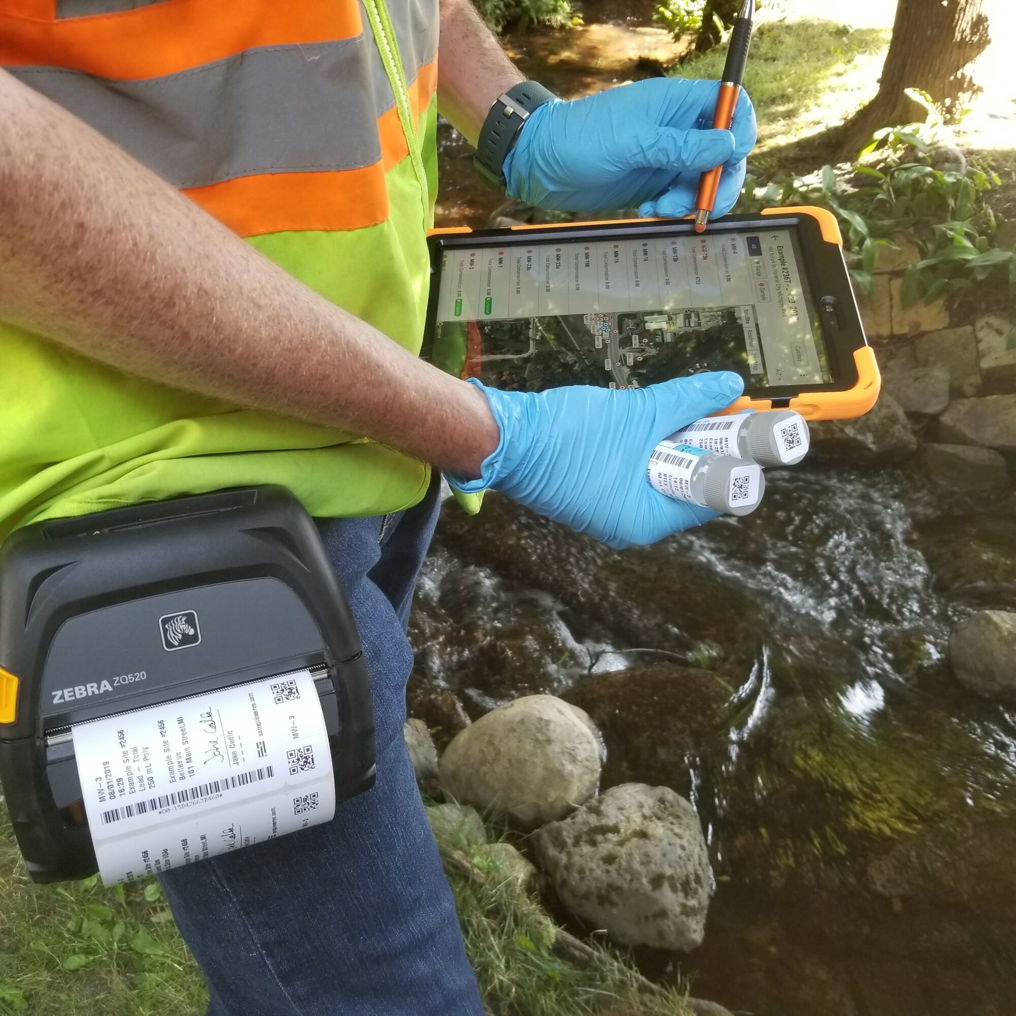 Person in safety vest and blue gloves holding a tablet and sampling equipment near a stream.