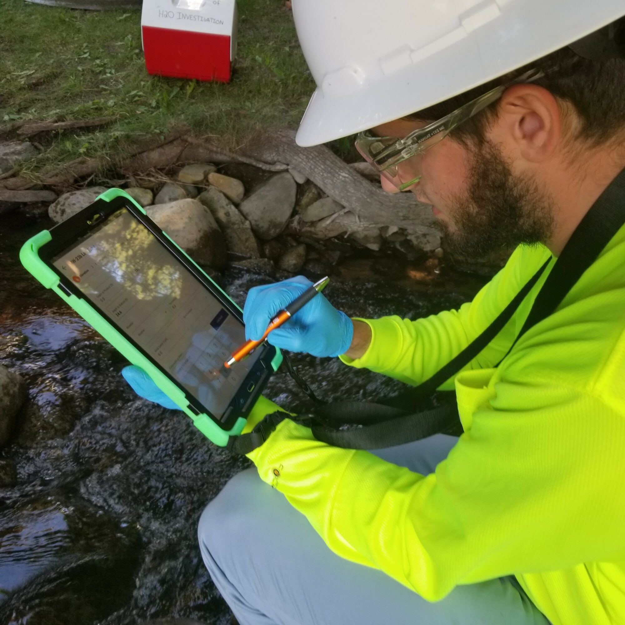 Person in safety gear using a tablet near a stream, holding a pen or stylus, outdoors.