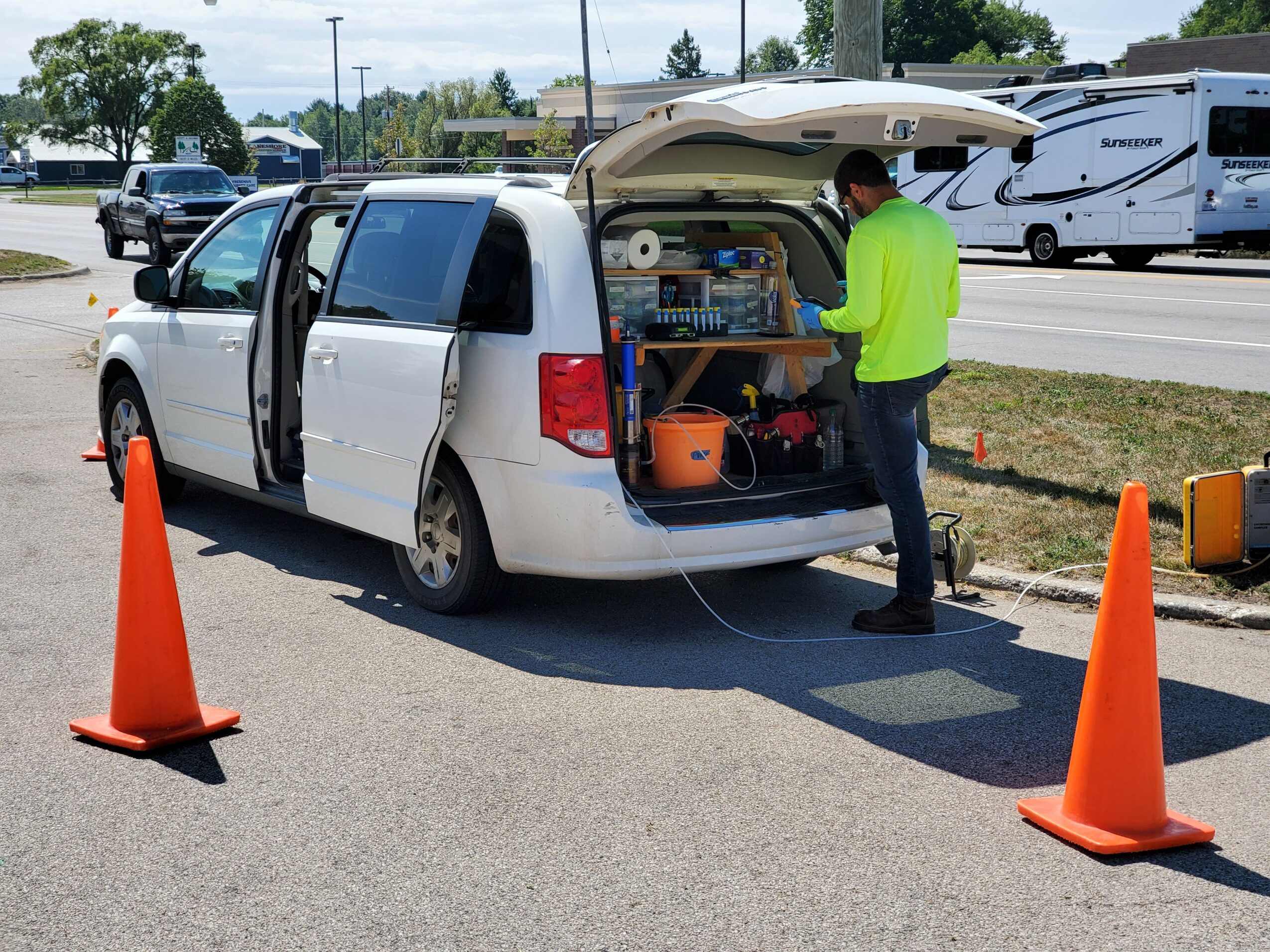 Person in neon yellow shirt loading equipment into open van trunk, orange cones on road, parked vehicles in background.