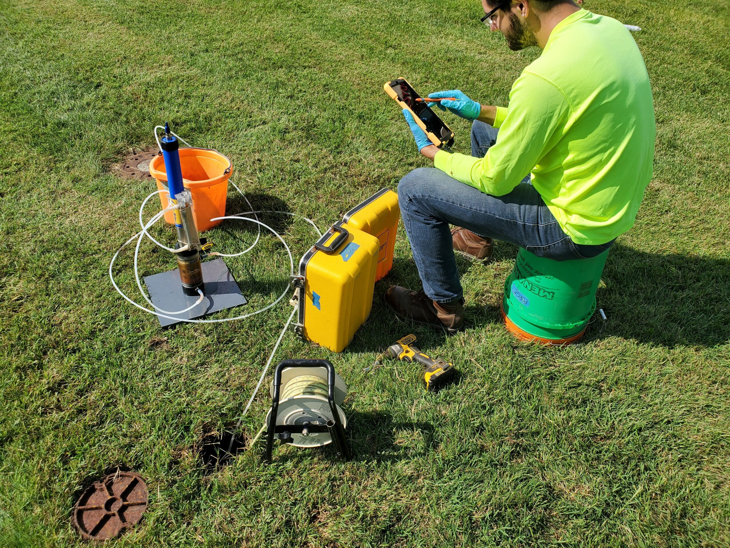 Person in neon green shirt and jeans sitting on a green bucket on grass, using equipment for soil testing.