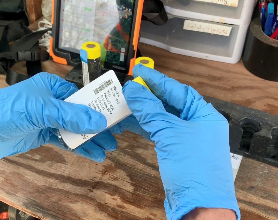 Person wearing blue gloves holding a small white object near a tablet and a black tool on a wooden surface.