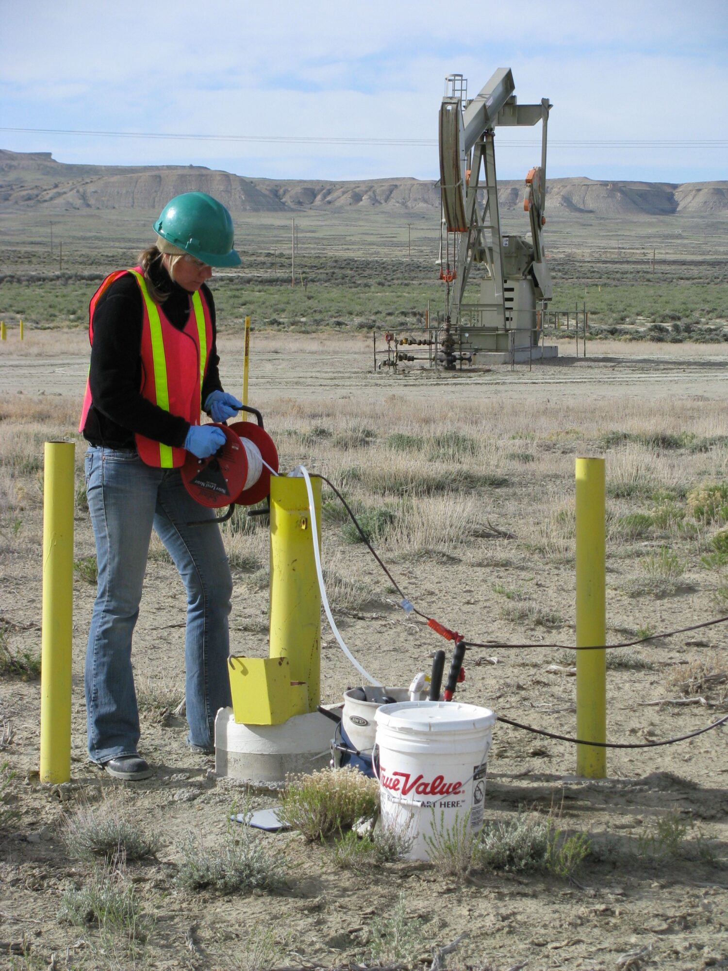 Person in safety gear working with equipment outdoors in a desert landscape with industrial structures in background.