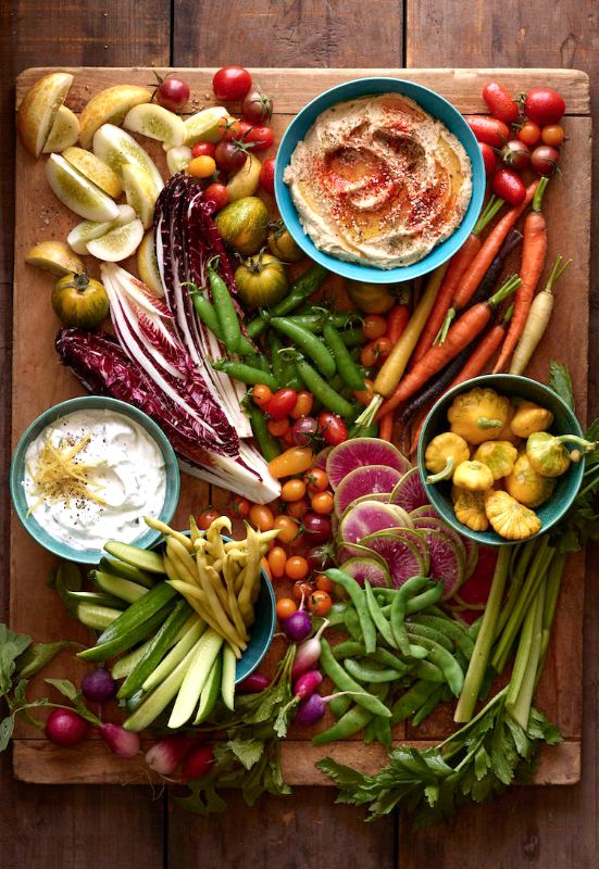 Assorted fresh vegetables and dips arranged on a wooden surface, including carrots, radishes, tomatoes, celery, and bowls of dips.