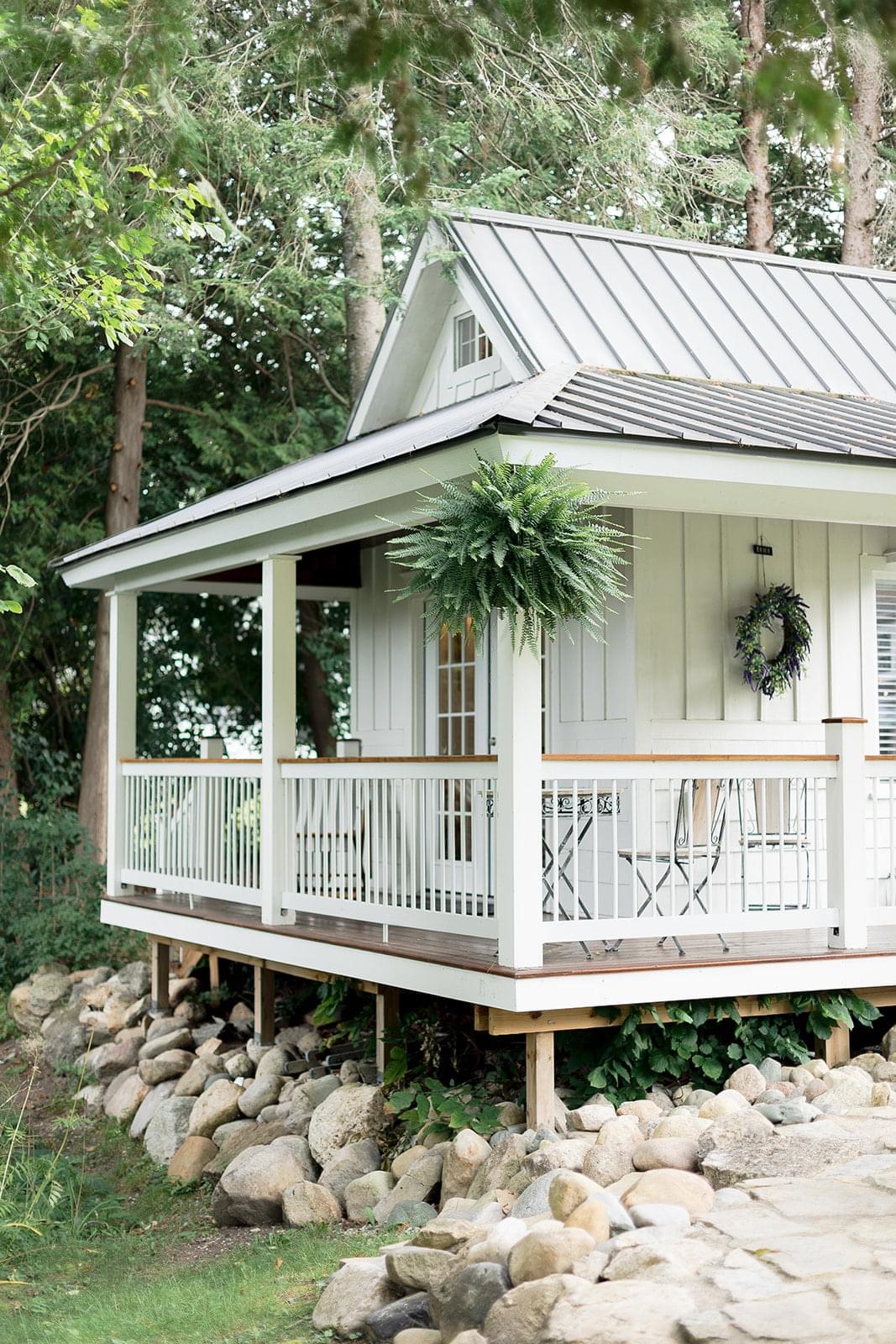 Porch of a house with white railing, potted plants, and outdoor furniture, surrounded by trees and rocks.