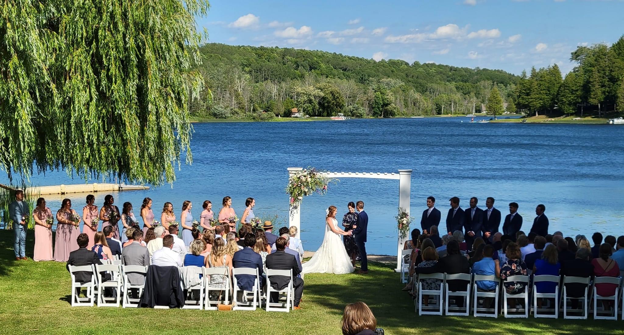 Wedding ceremony by a lake with guests seated, bride and groom at altar, scenic trees and water in background.