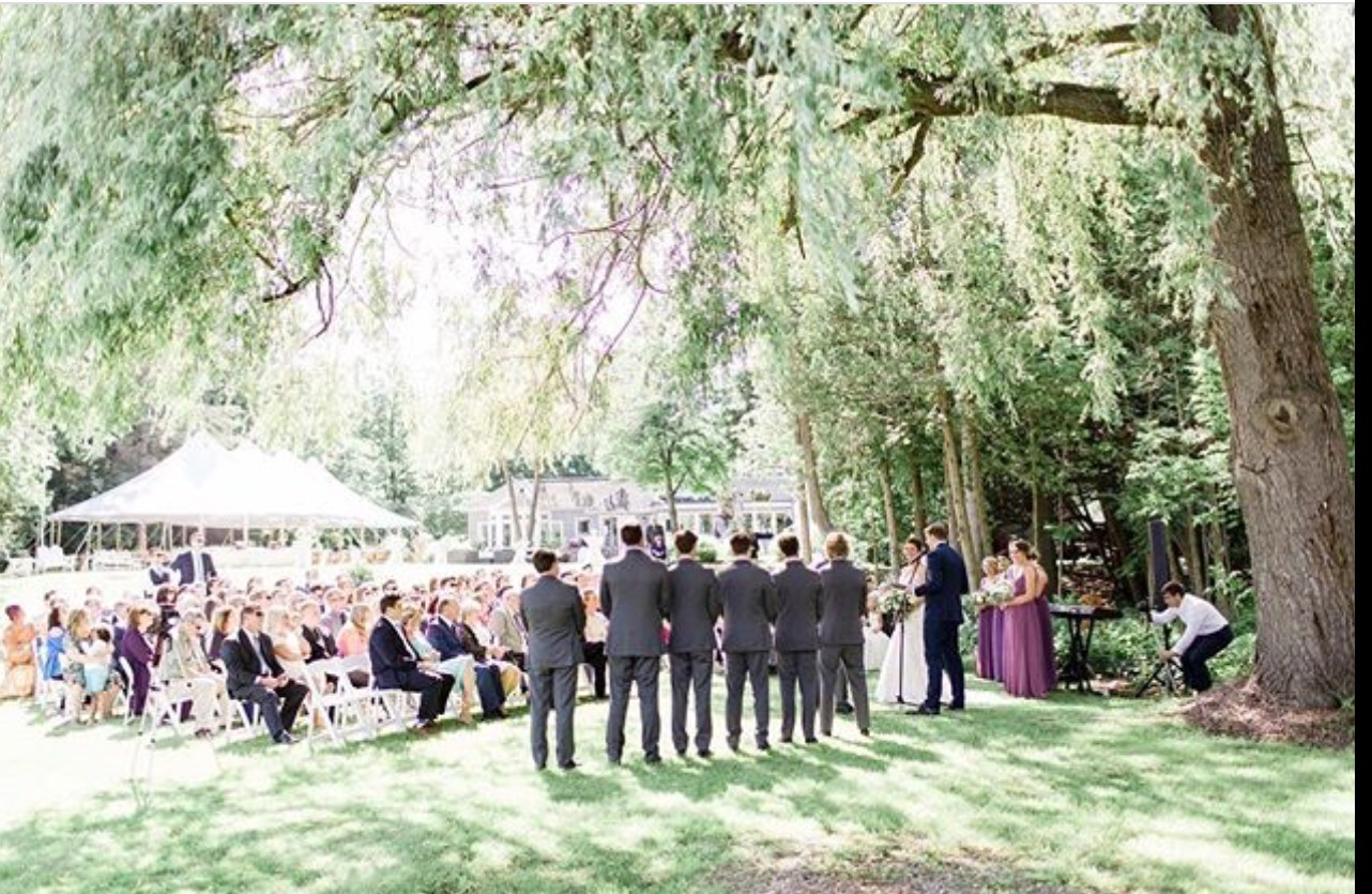 Group of people gathered outdoors under trees, some seated, some standing, with a tent in the background.