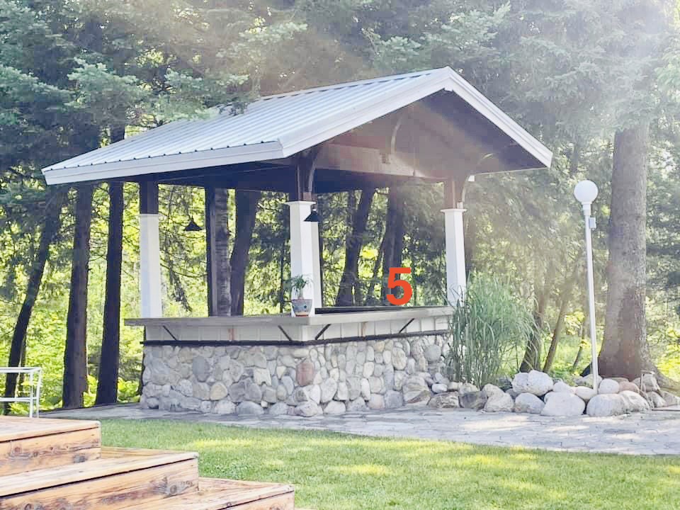 Stone structure with a metal roof, surrounded by trees and grass, with a red number 5 and a lamp post nearby.