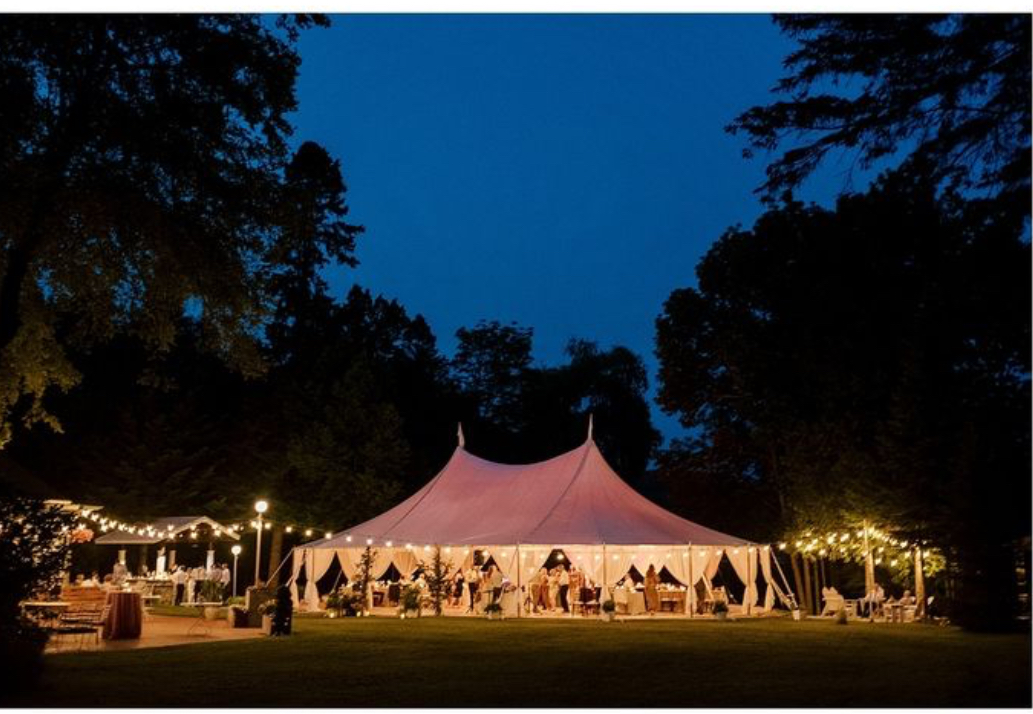 Large white tent illuminated at night, surrounded by trees and string lights, with a grassy area in front.