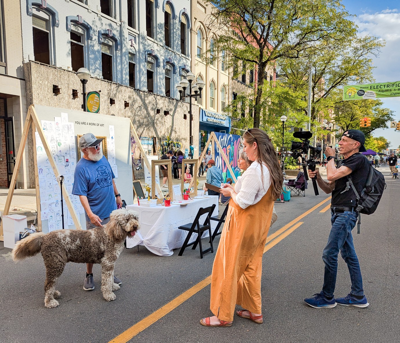 Person with a dog taking a photo of a woman at an outdoor event on a city street.