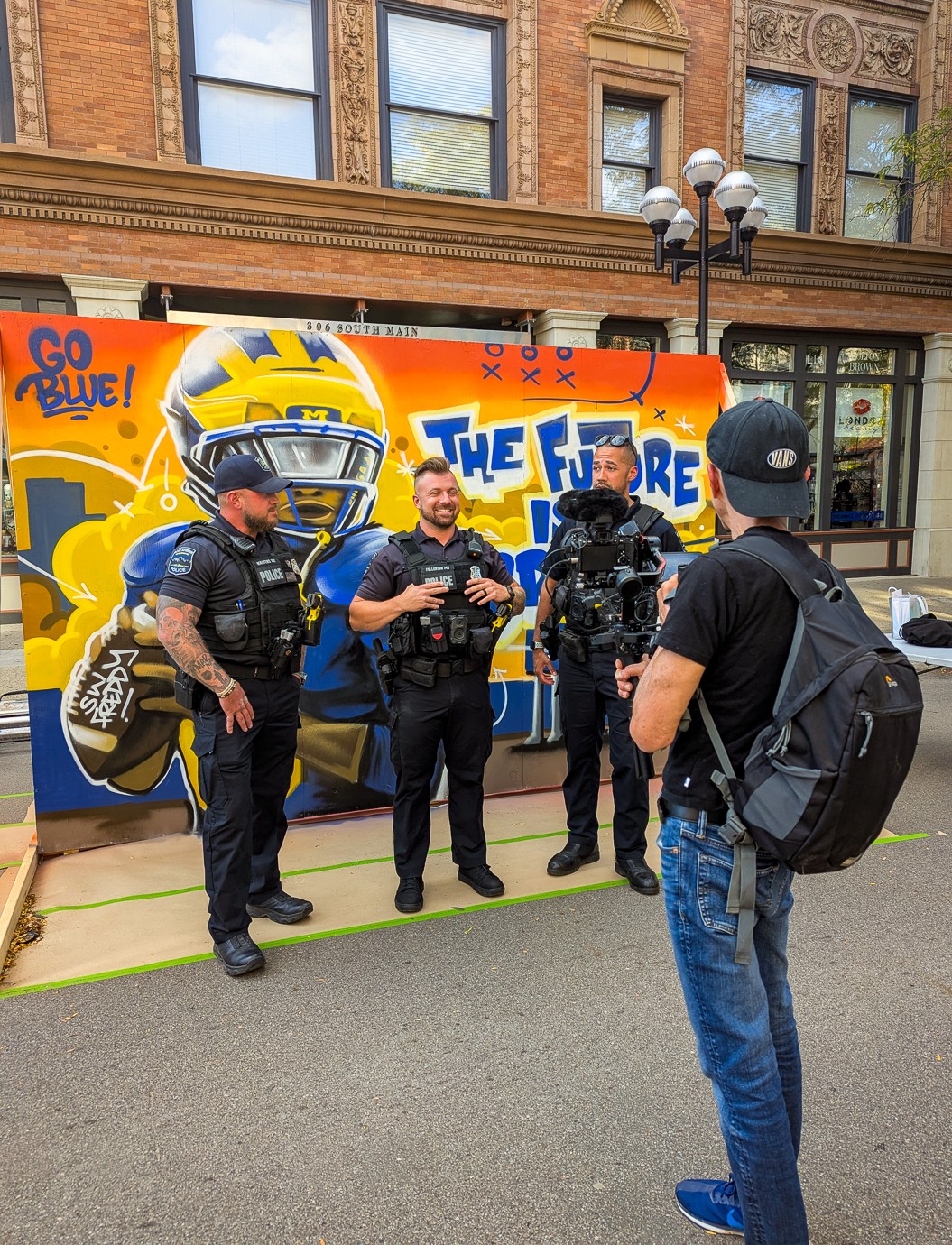 Three police officers and a man with a backpack talking in front of a colorful bus with a helmet graphic and text.