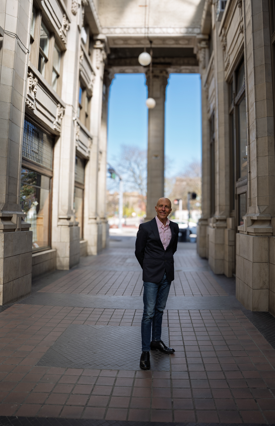 Man standing in a city street between tall historic buildings with columns and a blue sky background.