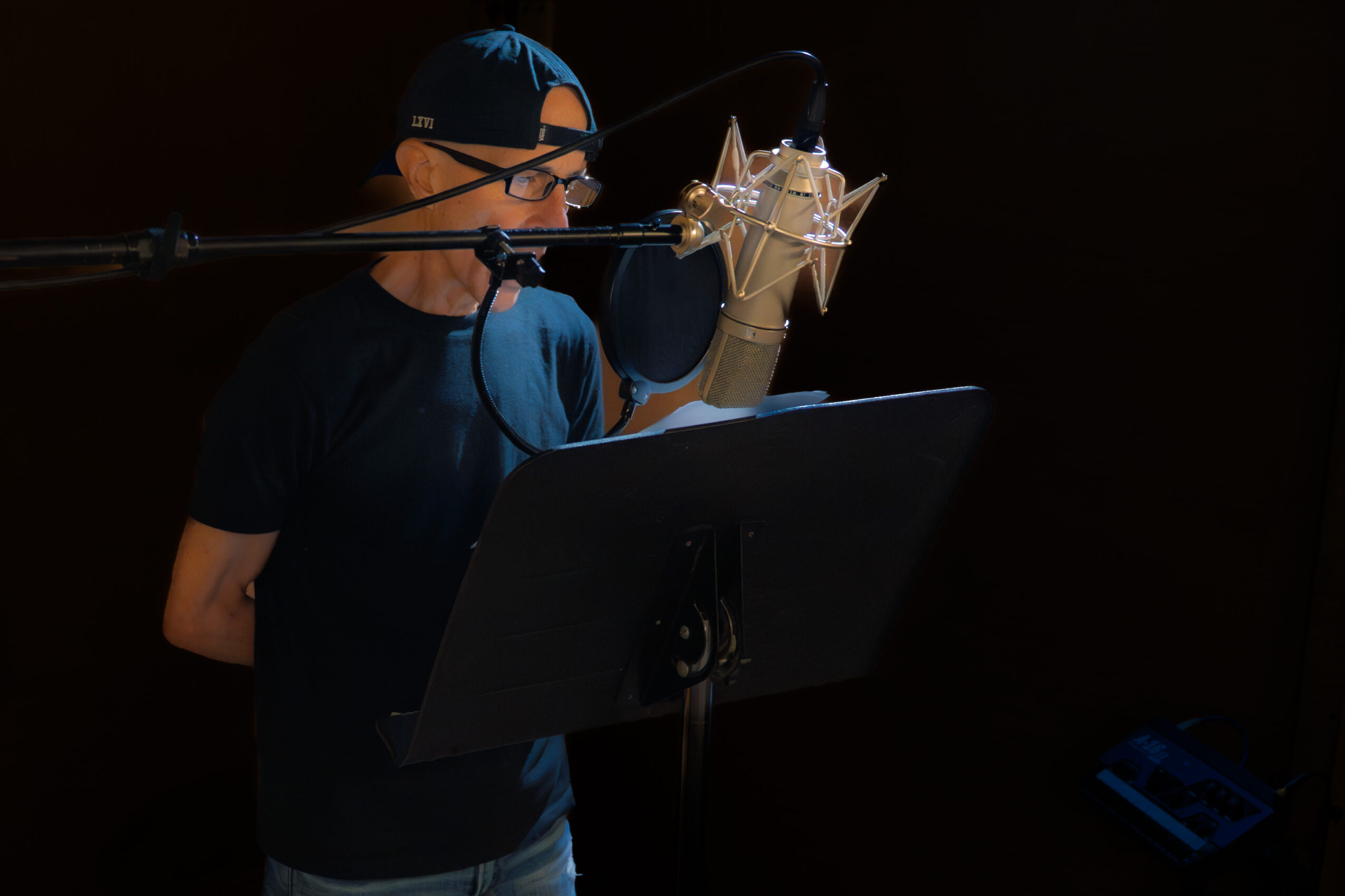 Person wearing glasses and a cap in a recording studio, speaking into a microphone with a pop filter, reading from a script.