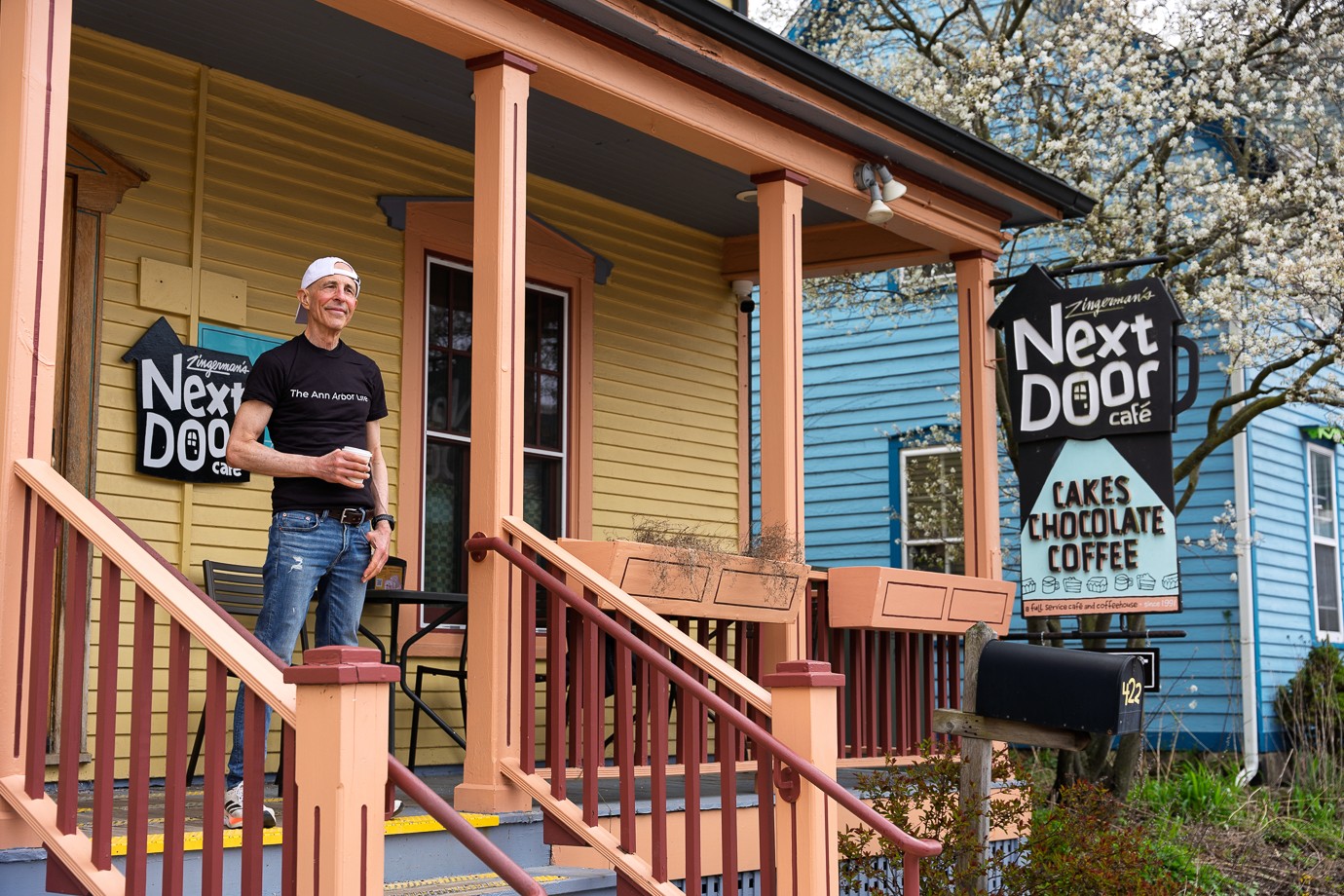 Person standing on porch holding a drink, next to signs advertising cakes, chocolate, and coffee, with houses in background.