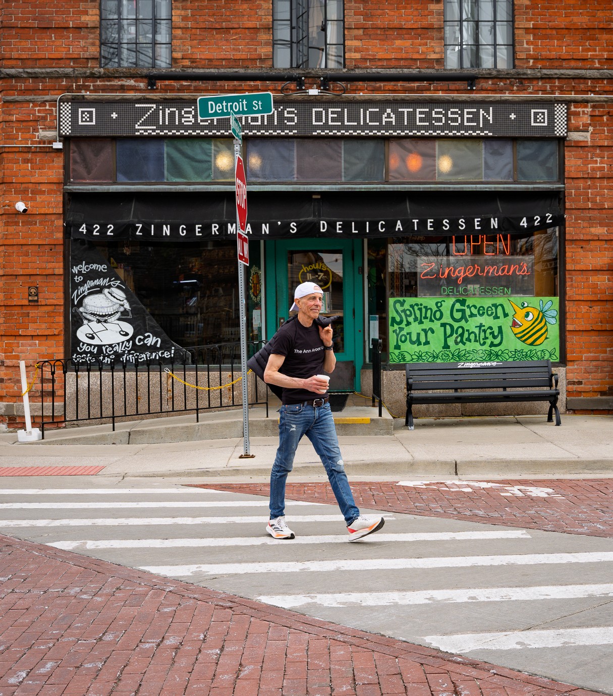 Person crossing street in front of Zingerman's Delicatessen storefront with green sign and bench, brick building background.