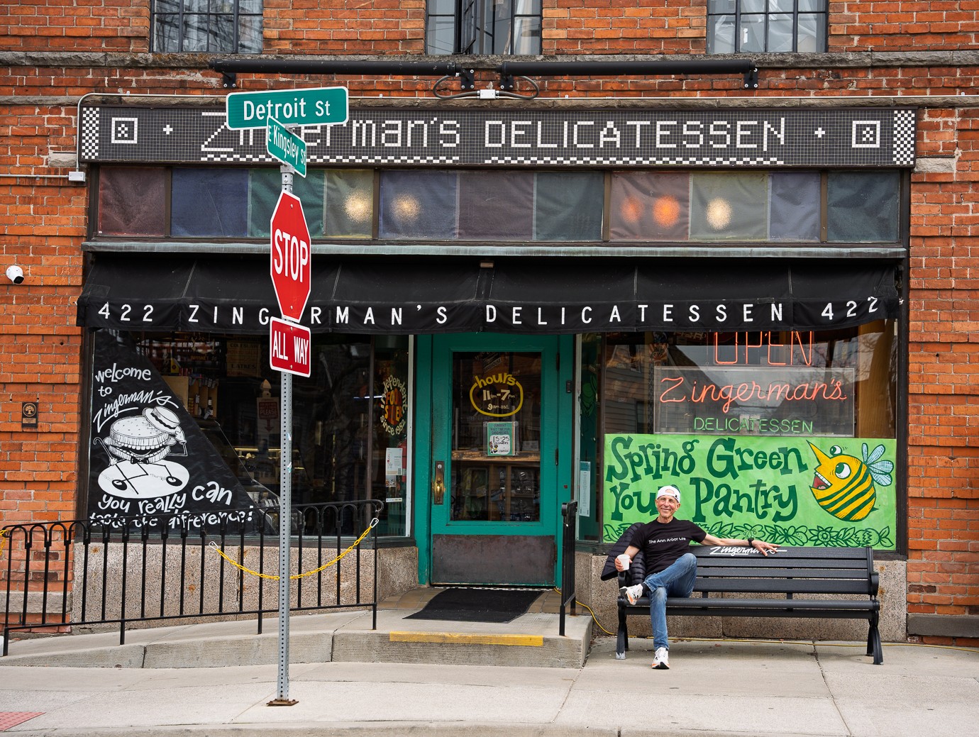Person sitting on a bench outside a delicatessen with a green sign and a bee illustration.