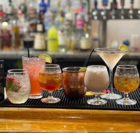 Seven colorful cocktails on a bar counter with blurred bottles in background.