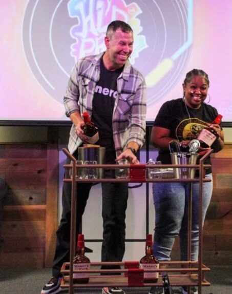 Two people standing behind a bar setup with glasses and bottles, smiling, in front of a screen with a logo.