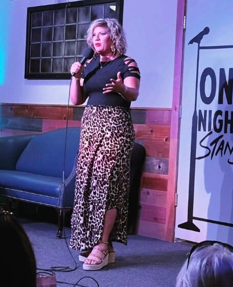 Woman with curly blonde hair holding a microphone on stage, wearing a black top and leopard print pants.