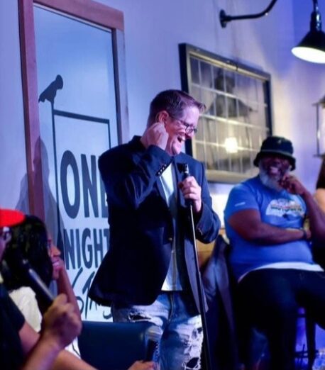 Man in blazer and glasses speaking into microphone on stage, with woman in hat and blue shirt seated nearby.