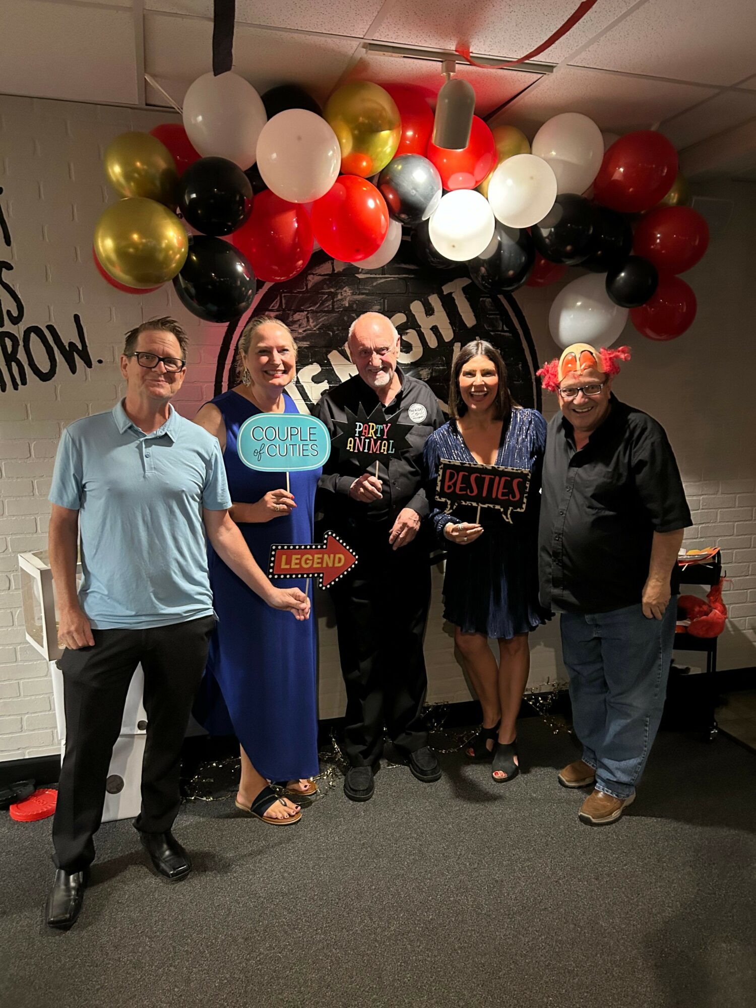 Five people standing in front of a balloon arch, smiling, holding signs, in a room with a decorated wall.