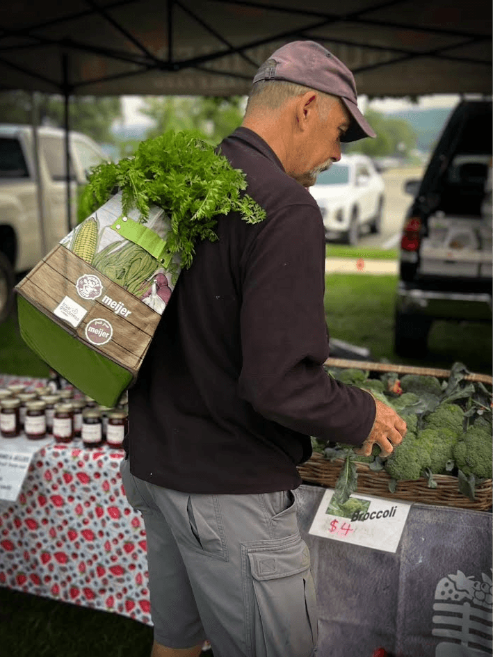 Man shopping at outdoor market, carrying a bag of leafy greens on his shoulder, browsing vegetables on table.