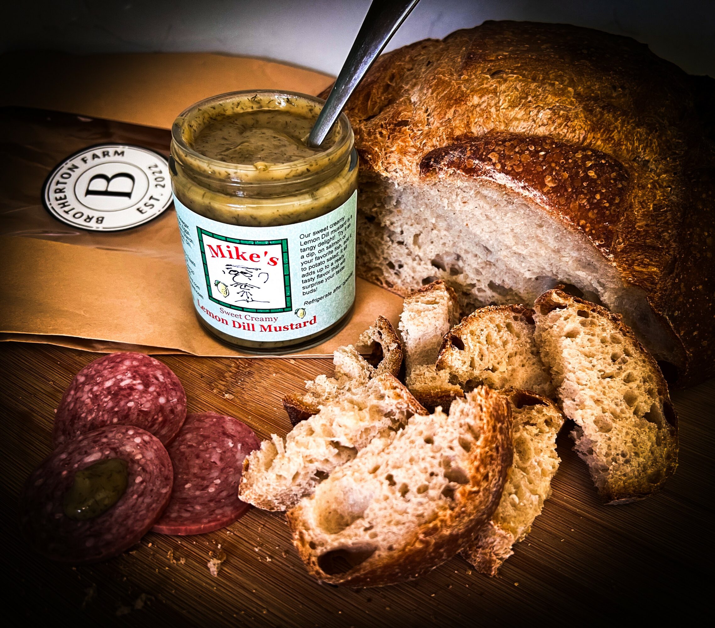 Open jar of spicy mustard, sliced bread, loaf of bread, salami slices, and bread slices on a wooden surface.