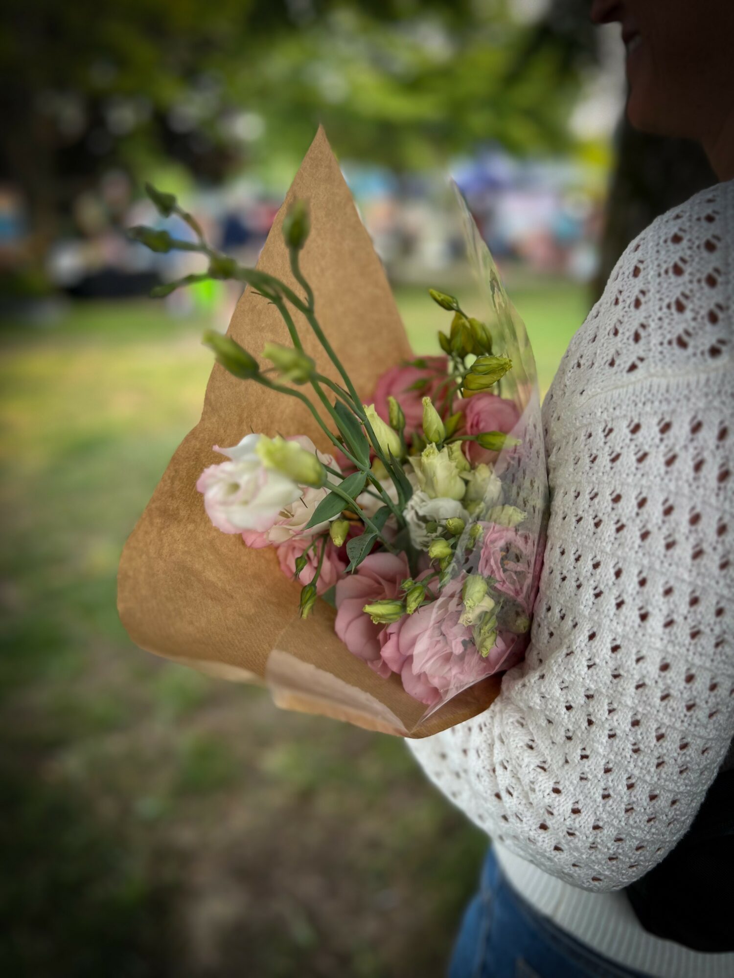 Person holding a bouquet of pink and white flowers wrapped in brown paper outdoors.
