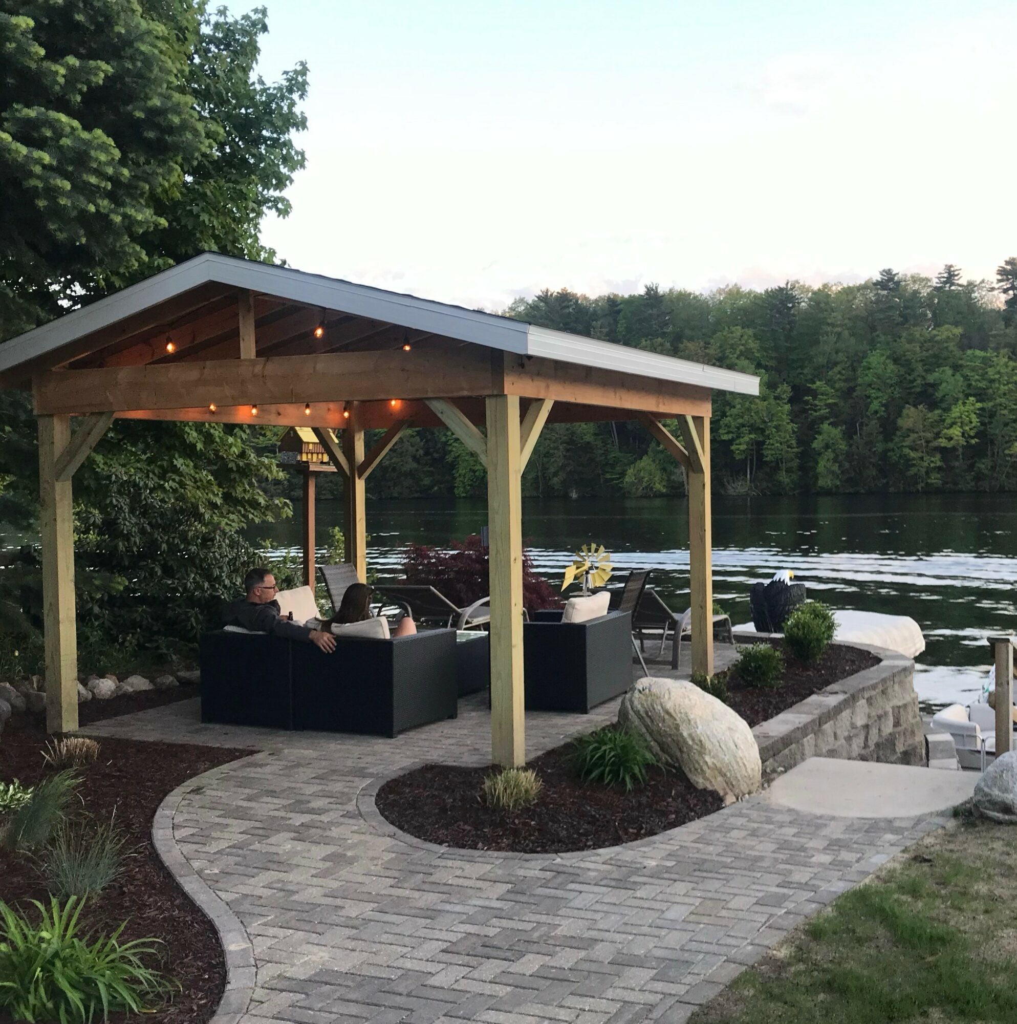 Outdoor seating area with a wooden pavilion by a river, surrounded by trees and a paved walkway.
