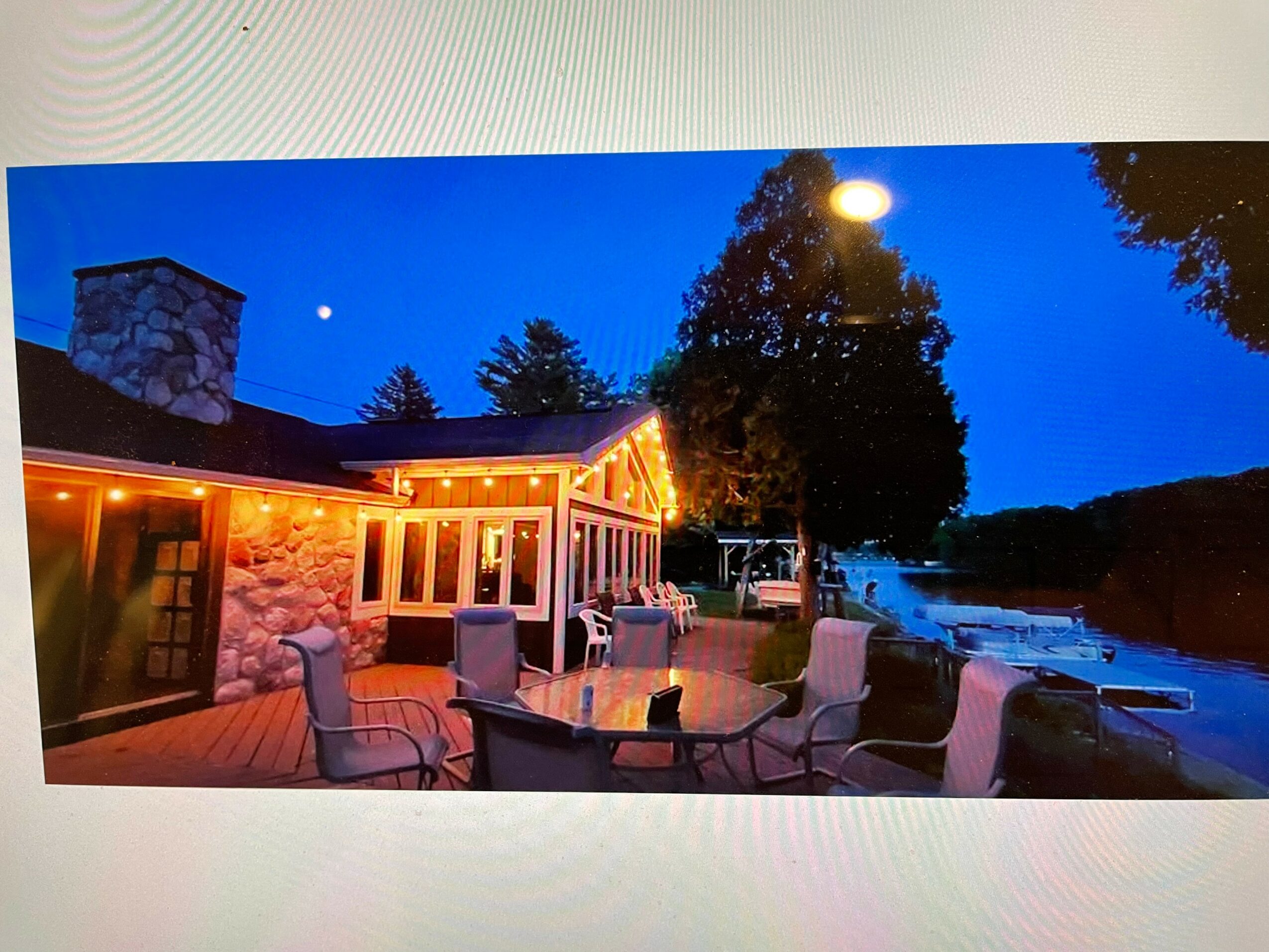 Outdoor patio with chairs and table, stone building, trees, river, evening sky with moon and star, string lights, illuminated scene