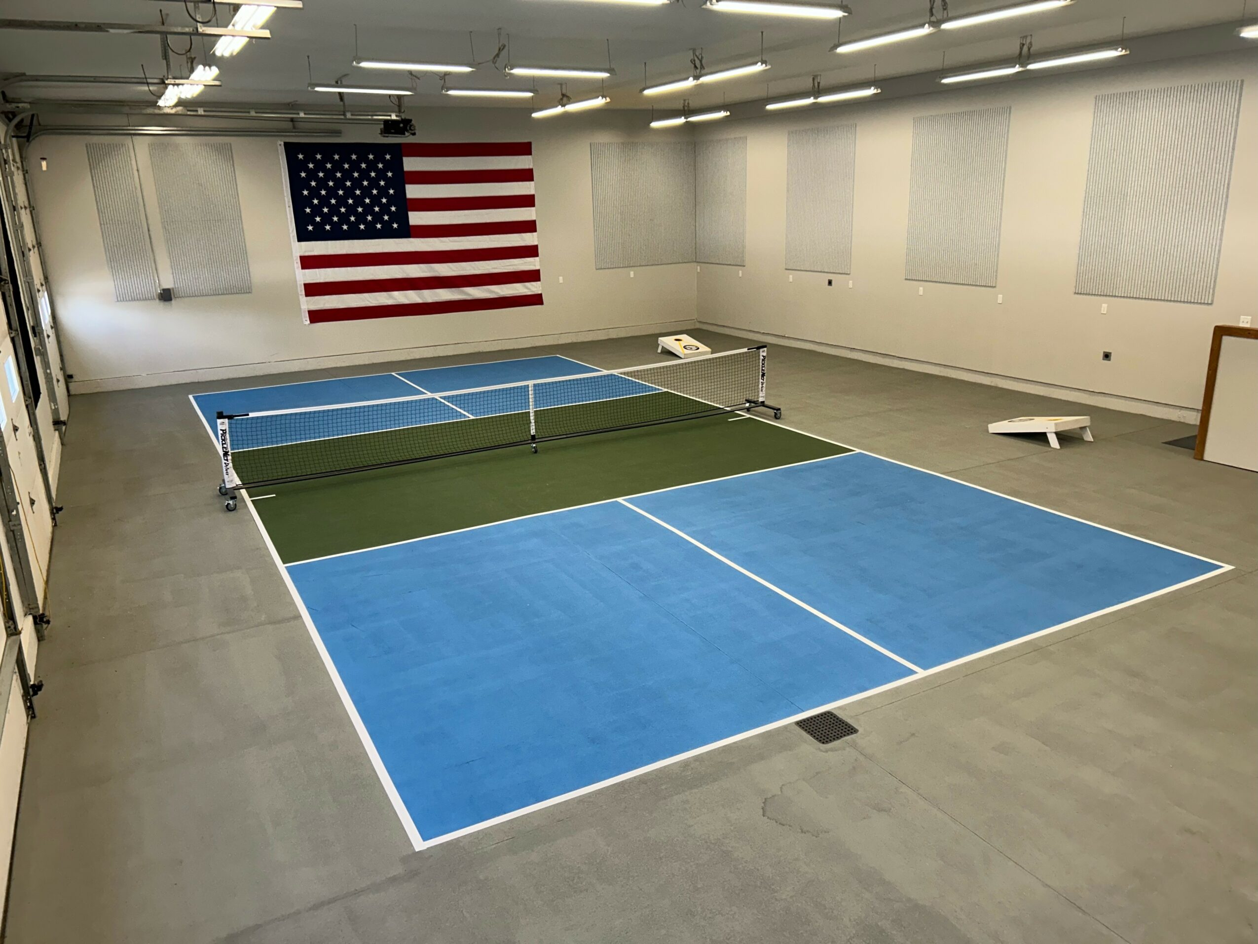 Indoor tennis court with blue and green surfaces, net, and American flag on the wall.