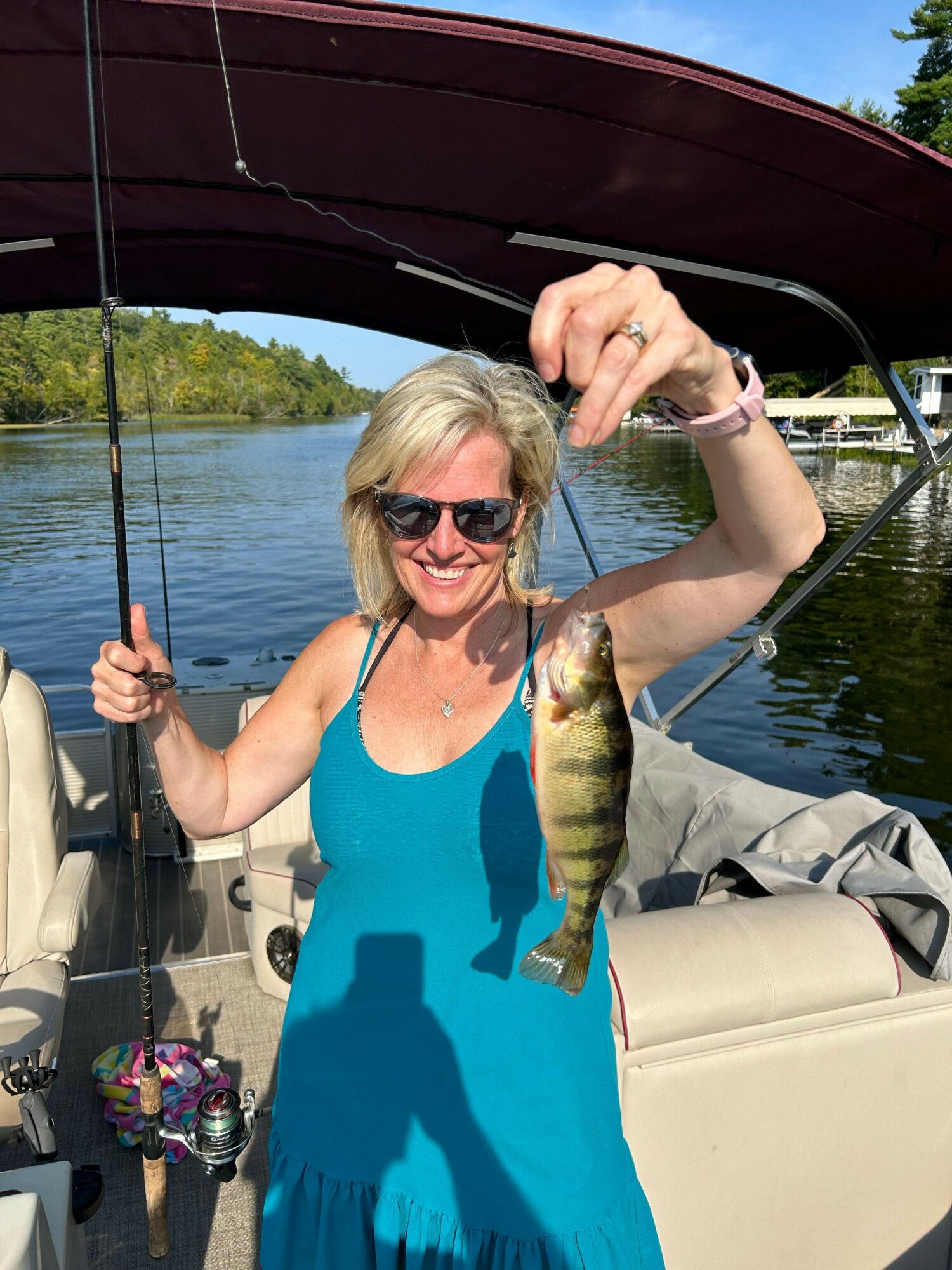 Woman with blonde hair and sunglasses holding a fish on a boat, smiling, with water and trees in background.