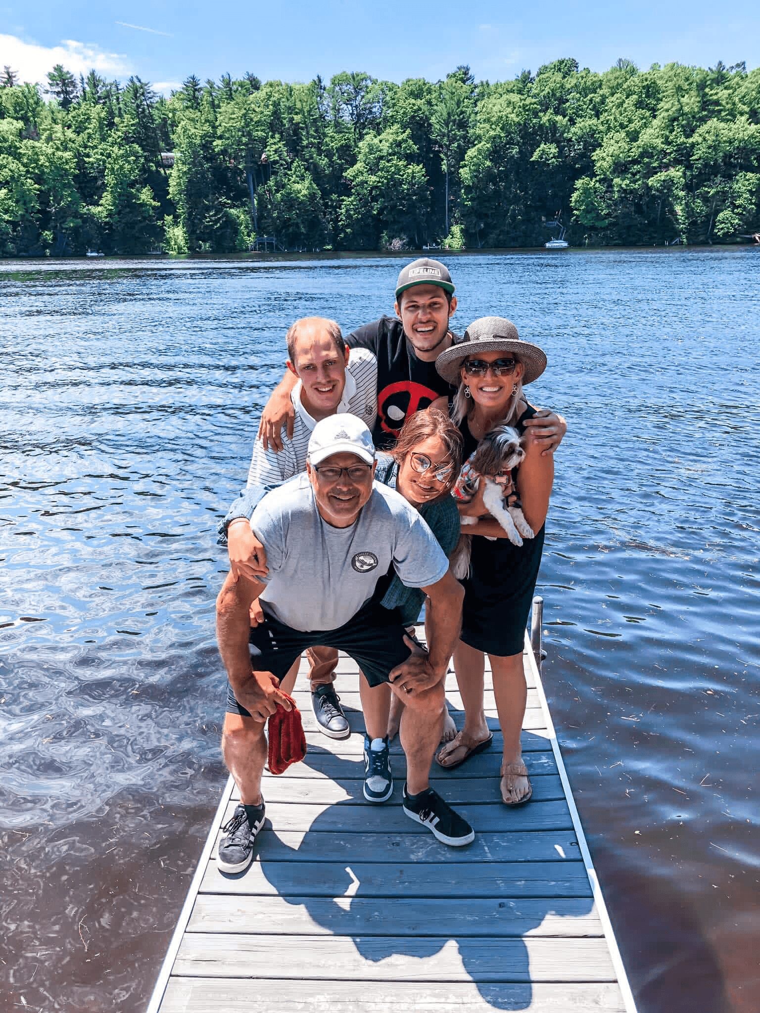 Group of five people standing on a dock by a lake, smiling, with trees in the background.
