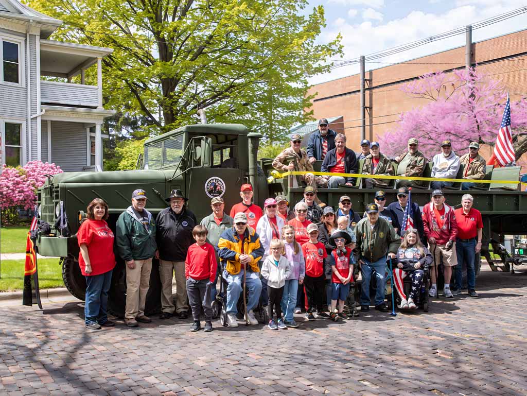 Group of children and adults standing in front of a green truck with a flatbed, some holding flags, in a park-like setting.