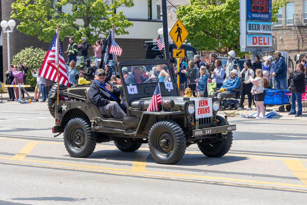 A small black vehicle with American flags, driving on a street during a parade with spectators and trees in background.