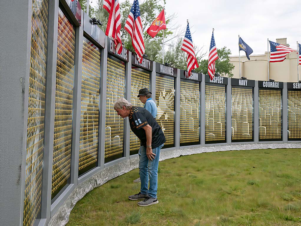 Two people look at a memorial wall with flags and names, one person leaning forward, the other standing behind.