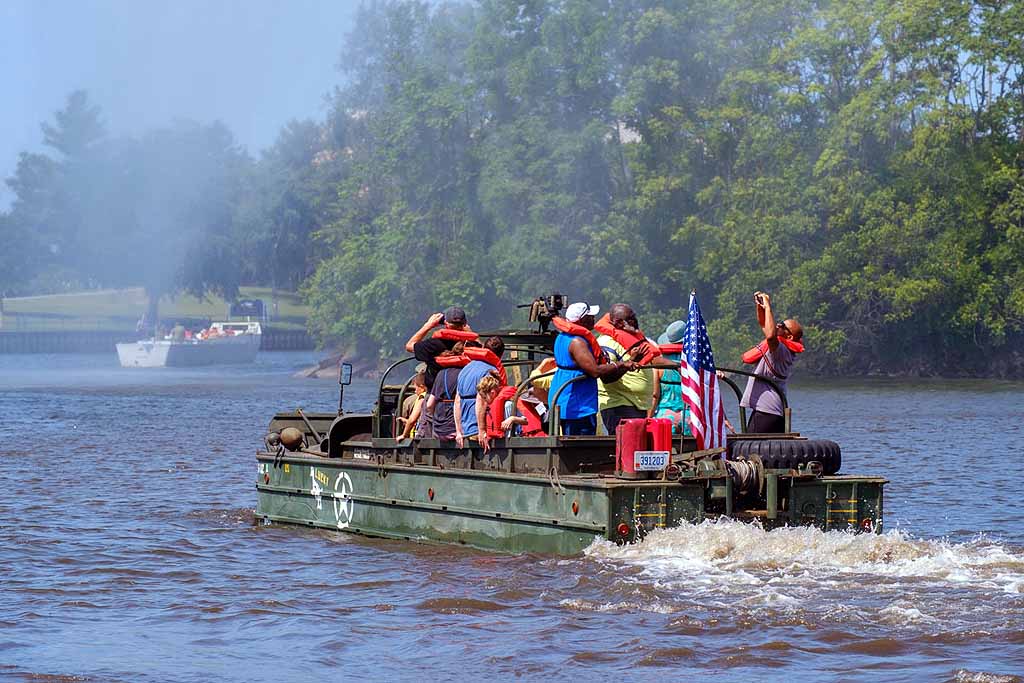 Group of people on a boat with American flags, wearing hats, on a river with trees in the background.