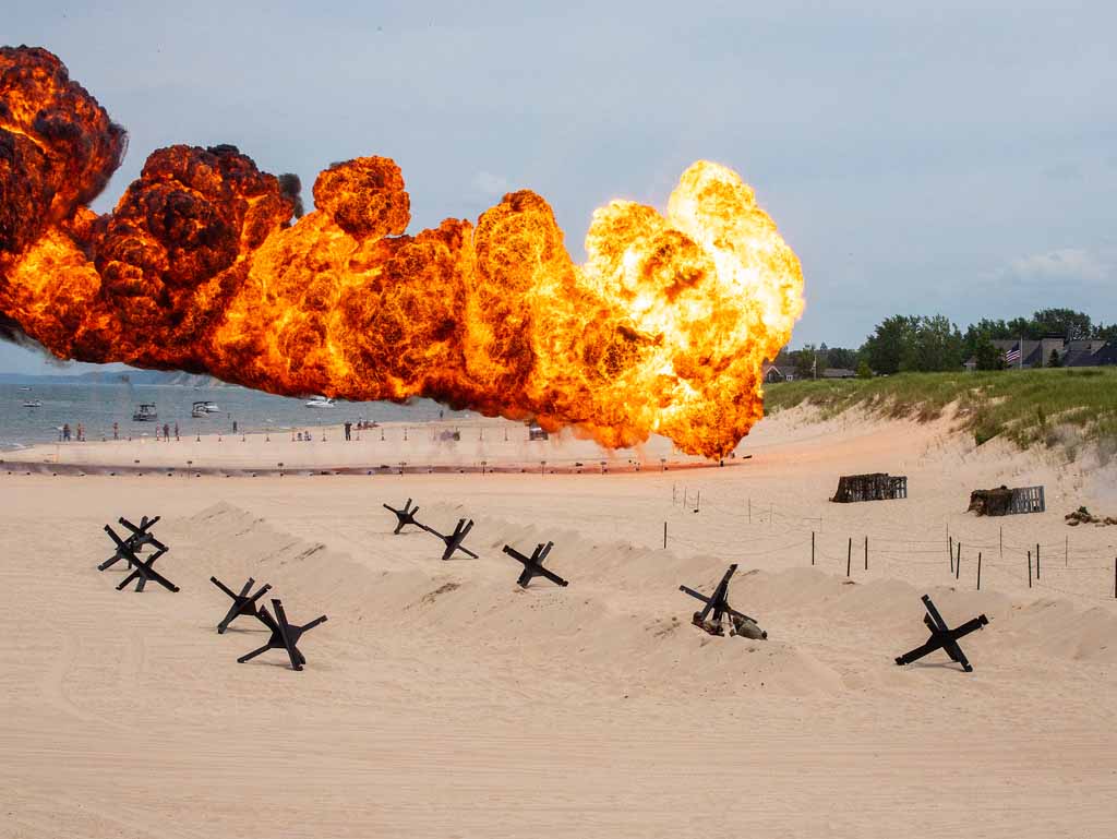 Explosion with fire and smoke on a beach, with sand and some objects in the foreground and trees in the background.