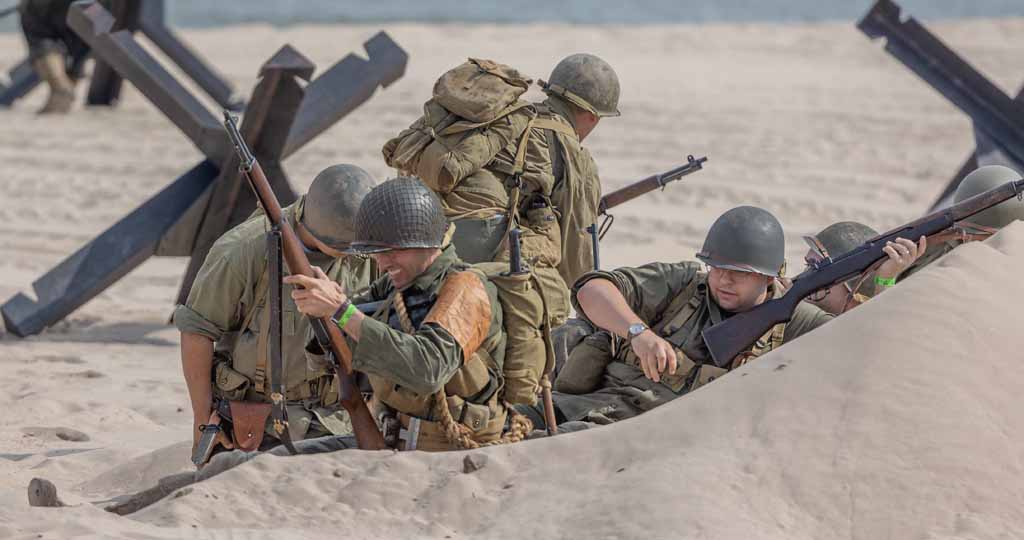 Group of soldiers in helmets and military gear lying in sand, holding rifles, with a large structure in the background.