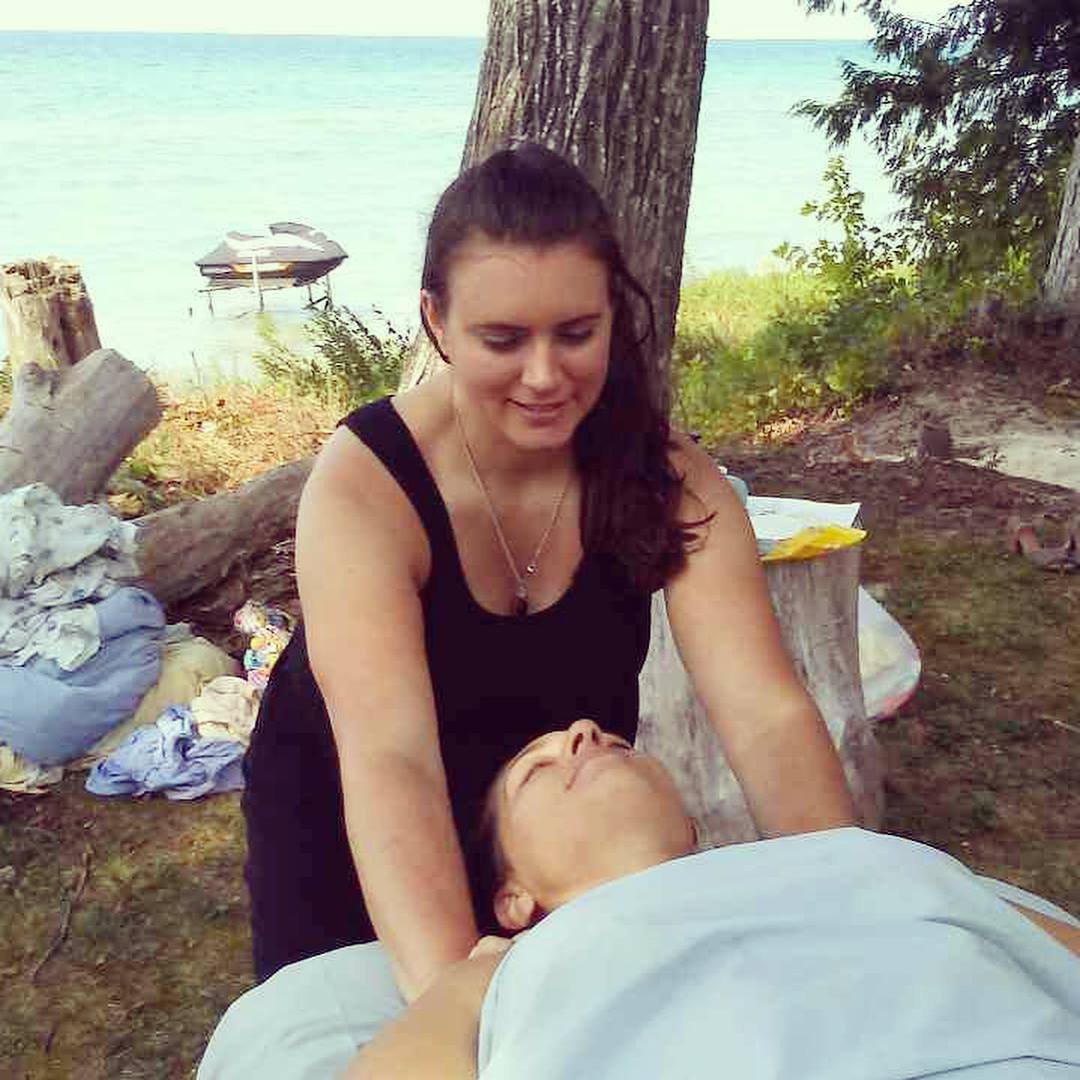 Woman in black dress providing first aid to a person lying on a blanket outdoors near water, trees, and logs.