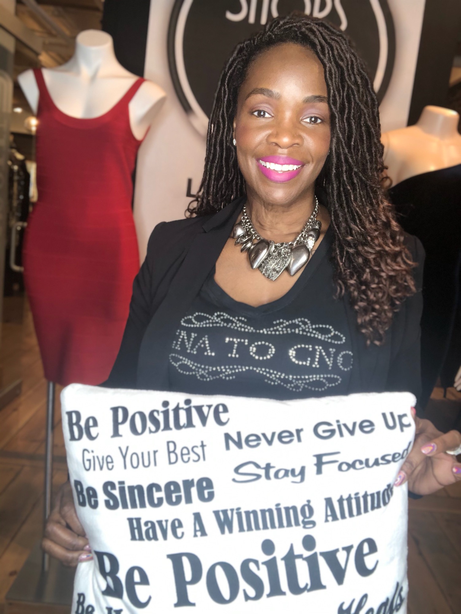 Woman holding a sign, smiling, wearing a black shirt and necklace, with mannequins and a sign in the background.