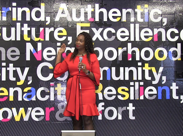 Woman in red dress speaking into microphone in front of colorful text background.