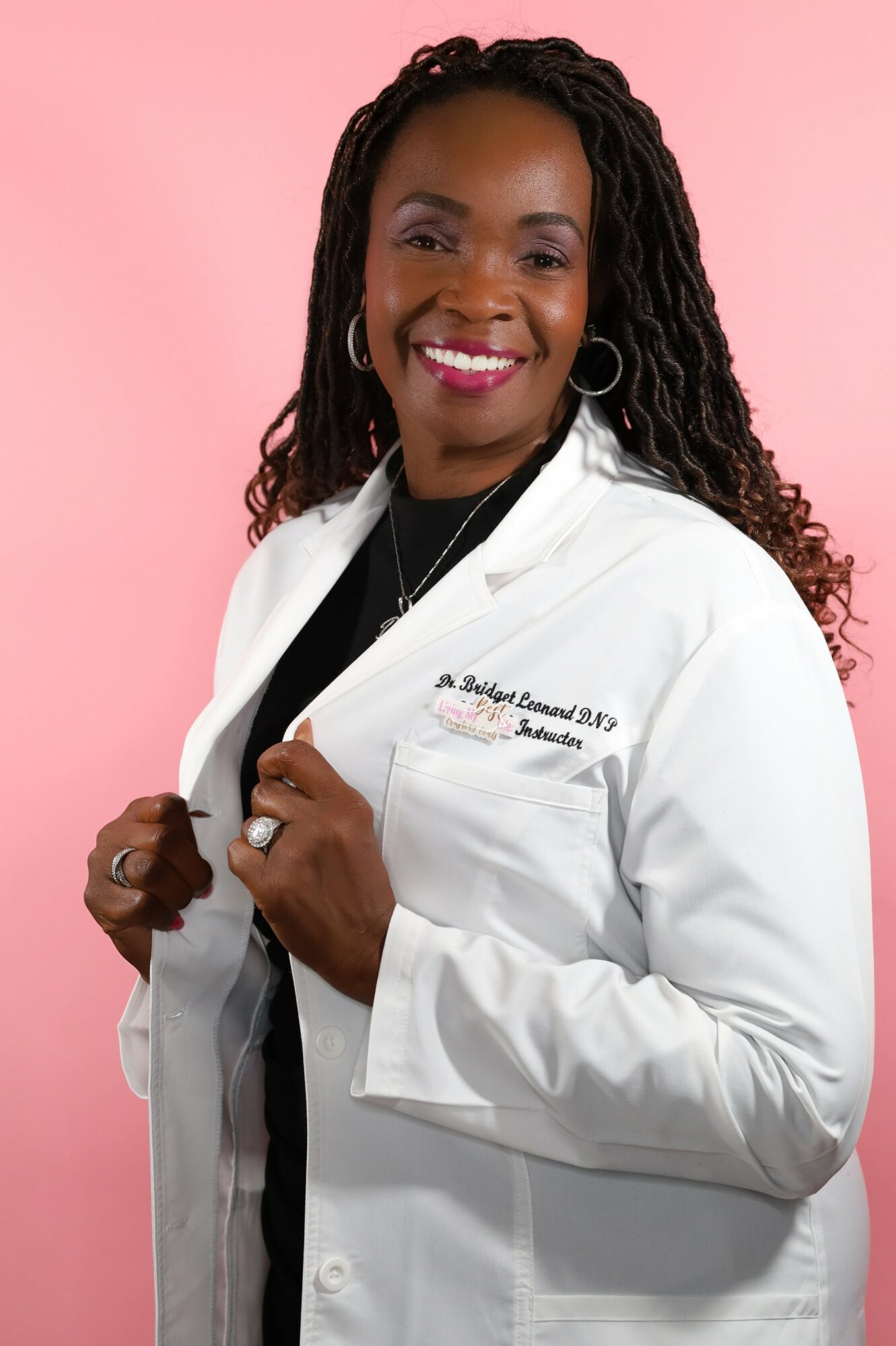 Woman in white lab coat smiling, holding lapels, pink background.