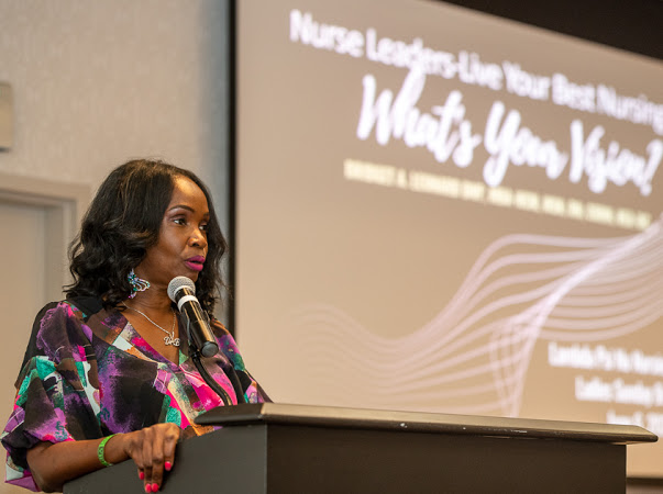 Woman speaking at a podium with a presentation screen behind her, wearing a colorful patterned top.